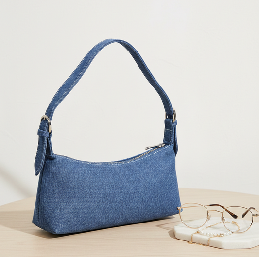 Blue denim shoulder bag with zipper on table next to gold glasses and pearl chain