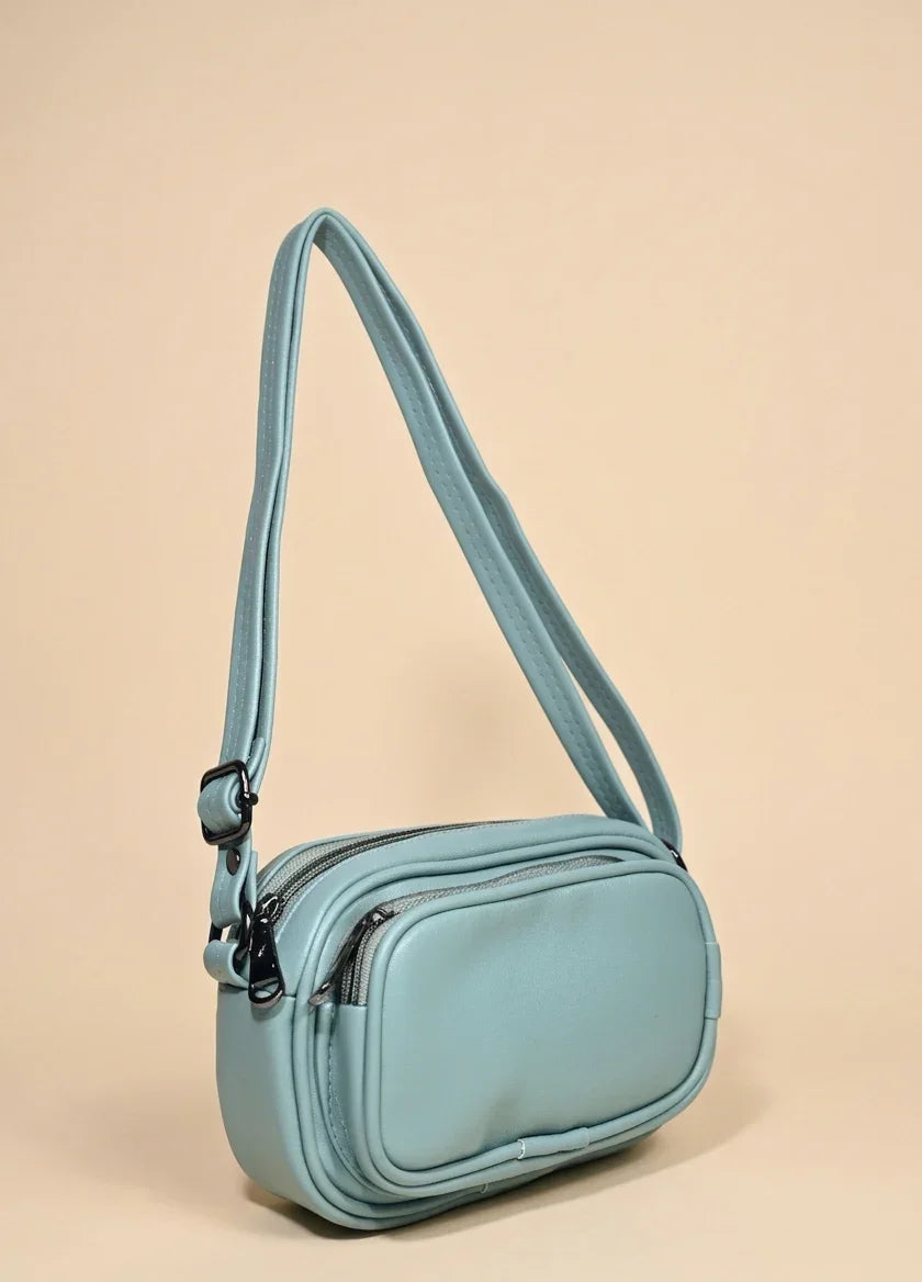 Light blue leather crossbody handbag with double zippers and adjustable strap on beige background